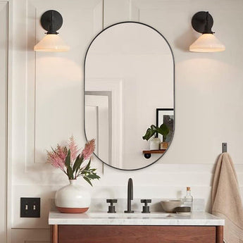 Black oval wall mirror with rounded arch frame above a white countertop and vase.