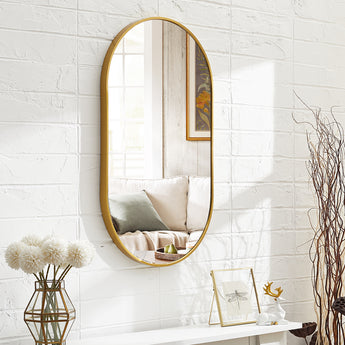 Gold oval wall mirror with thin frame mounted on a white brick wall above a bench.