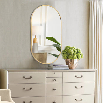 Gold oval wall mirror with slim frame above a white dresser and potted plant.