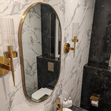 Gold oval wall mirror with slim metal frame reflecting a marble bathroom vanity.