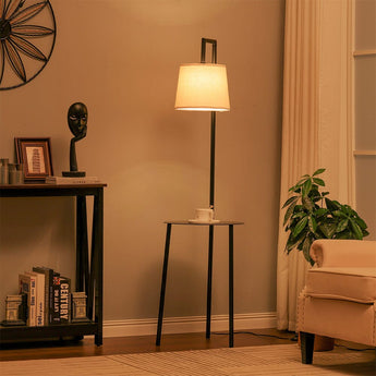 Black floor lamp with linen lampshade and round metal tray table on three legs.